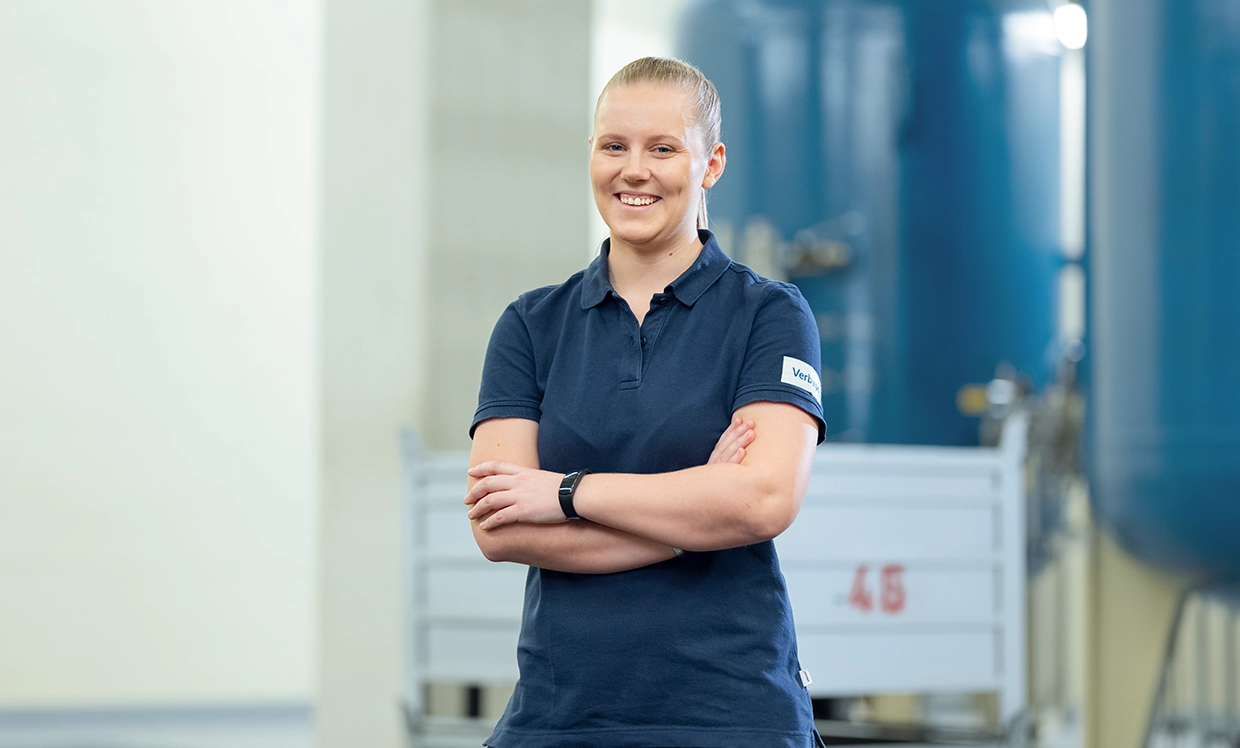 Eine junge technische Mitarbeiterin in dunkelblauem T-shirt lächelt ins Bild und steht mit verschränkten Armen in einer Halle