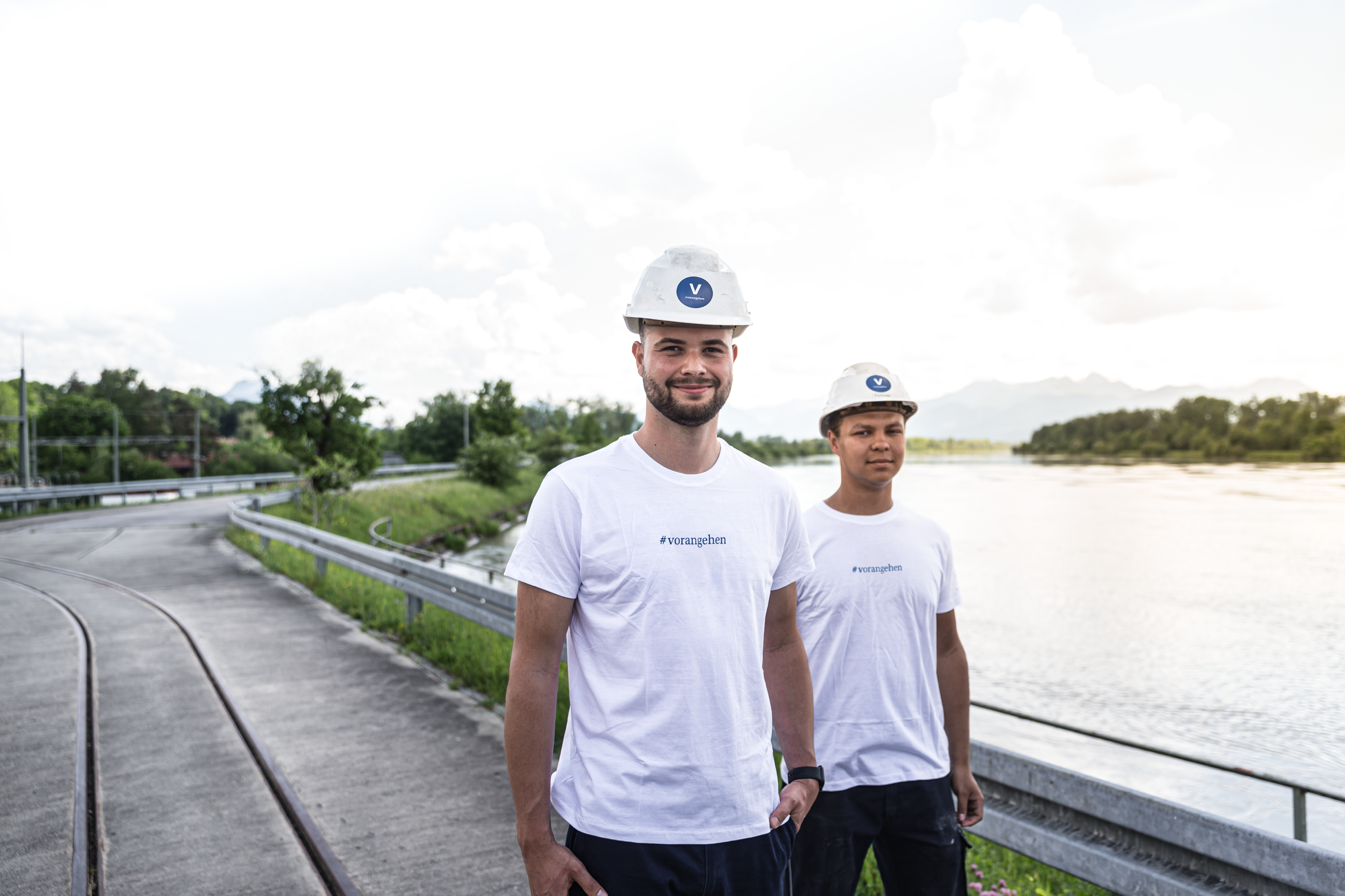 Am rechten Bildrand befinden sich zwei Lehrlinge in weißen T-Shirts und Helm. Im Hintergrund sehen wir die Donau, Bäume, Schienen und eine kleine Straße.