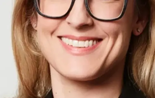 Domenika Koller has blonde hair that falls to her shoulders. She wears distinctive black glasses and a black blazer with delicate gold jewellery. She smiles sympathetically into the camera.