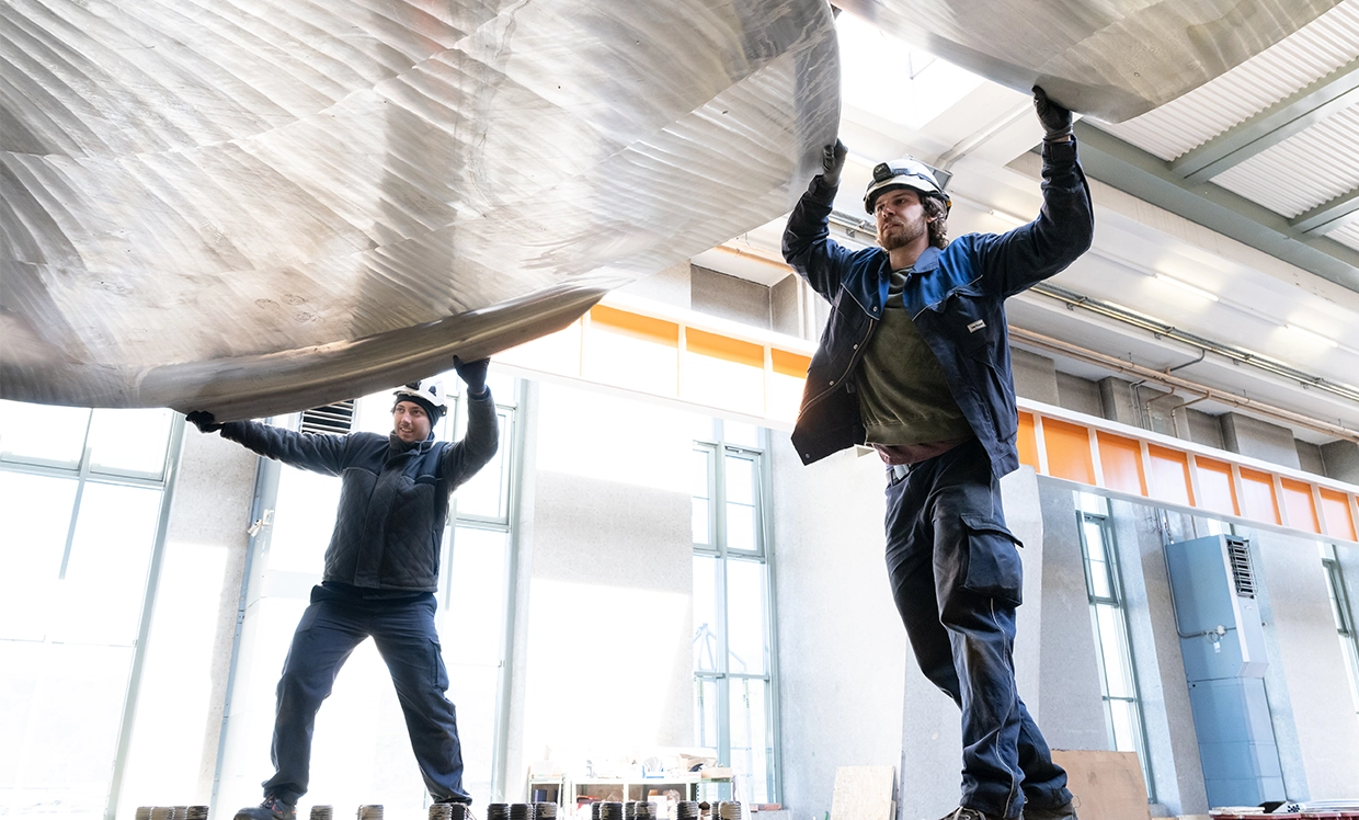 Wir sehen zwei VERBUND-Mitarbeiter bei der Arbeit an einer Turbine. Beide tragen Arbeitskleidung und Helme. Im Hintergrund sieht man das Wasserkraftwerk.