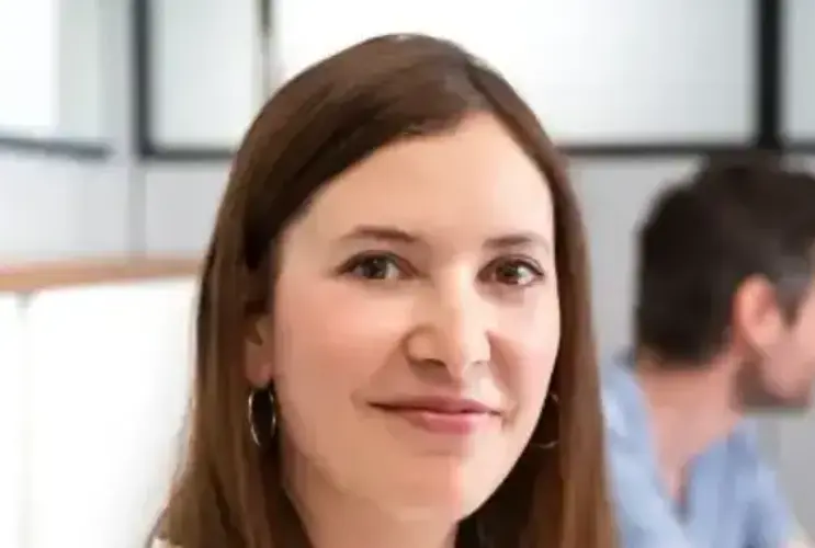 In einem modernen Büroraum sitzt eine junge Mitarbeiterin von VERBUND mit dunkelblonden glatten Haaren. Sie lächelt in die Kamera. Rechts von ihr sind im Hintergrund 2 männliche Kollegen zu sehen, die sich unterhalten.