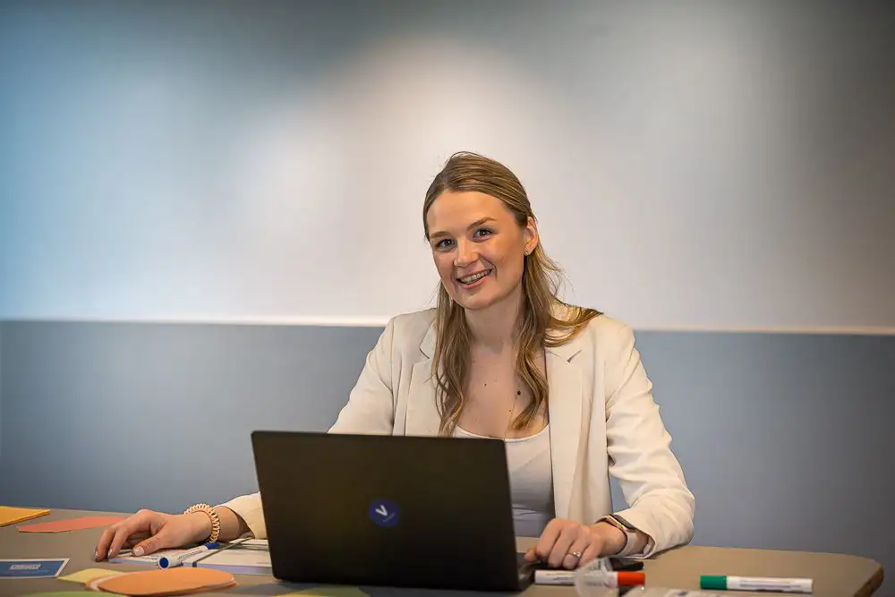 Eine VERBUND-Mitarbeiterin mit langen blonden Haaren und braunen Augen sitzt in einem Meetingraum. Sie sitzt vor ihrem Laptop und blickt lächelnd in die Kamera.