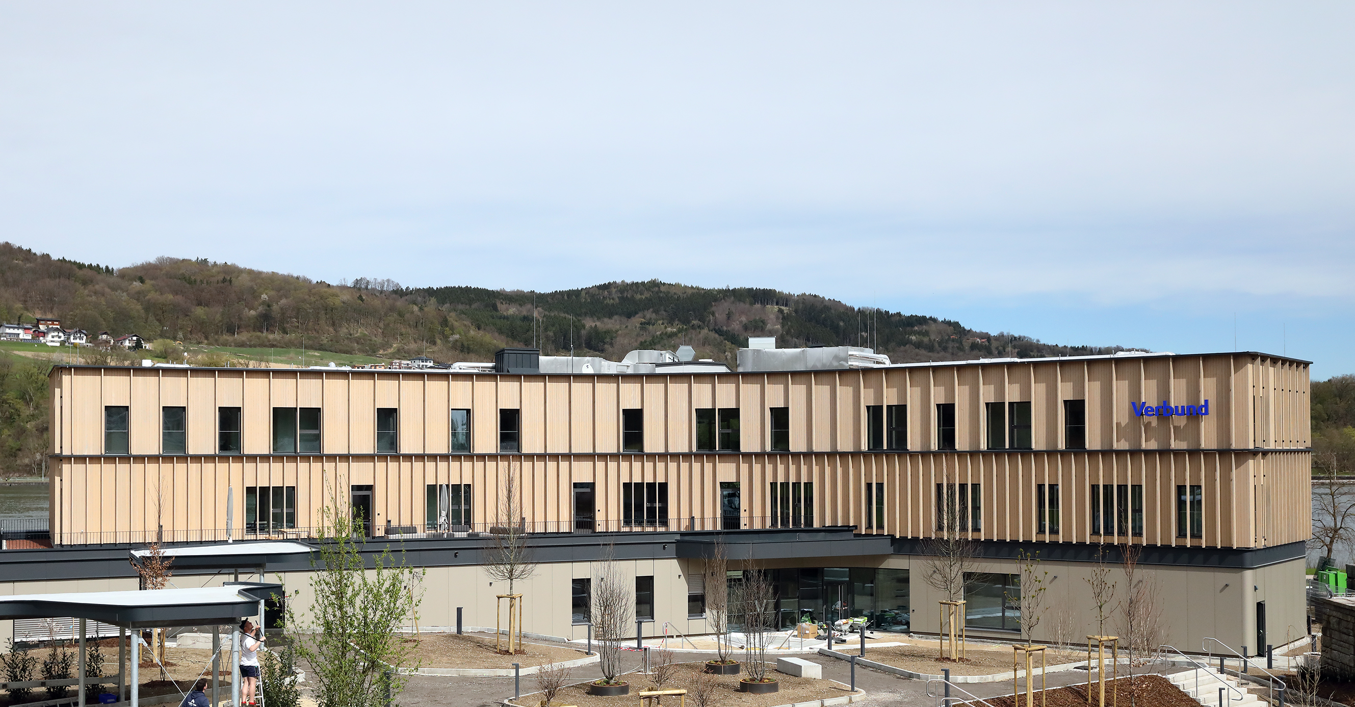 Wir blicken auf das neue Lehrlingsheim im Ybbs. Die Fasade ist beigefarben und blaue VERBUND-Schriftzug ist gut sichtbar. Das Gebäude befindet sich im ländlichen Raum. Im Hintergrund sieht man grüne Hügel.