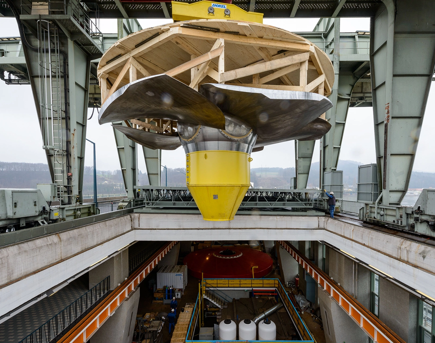 Wir beobachten zahlreiche VERBUND-Mitarbeiter.innen dabei, wie eine neue Turbine in ein Wasserkraftwerk eingebaut wird. Gerade eben wir die Turbine via Kran ins Innere des Kraftwerks abgeseilt.