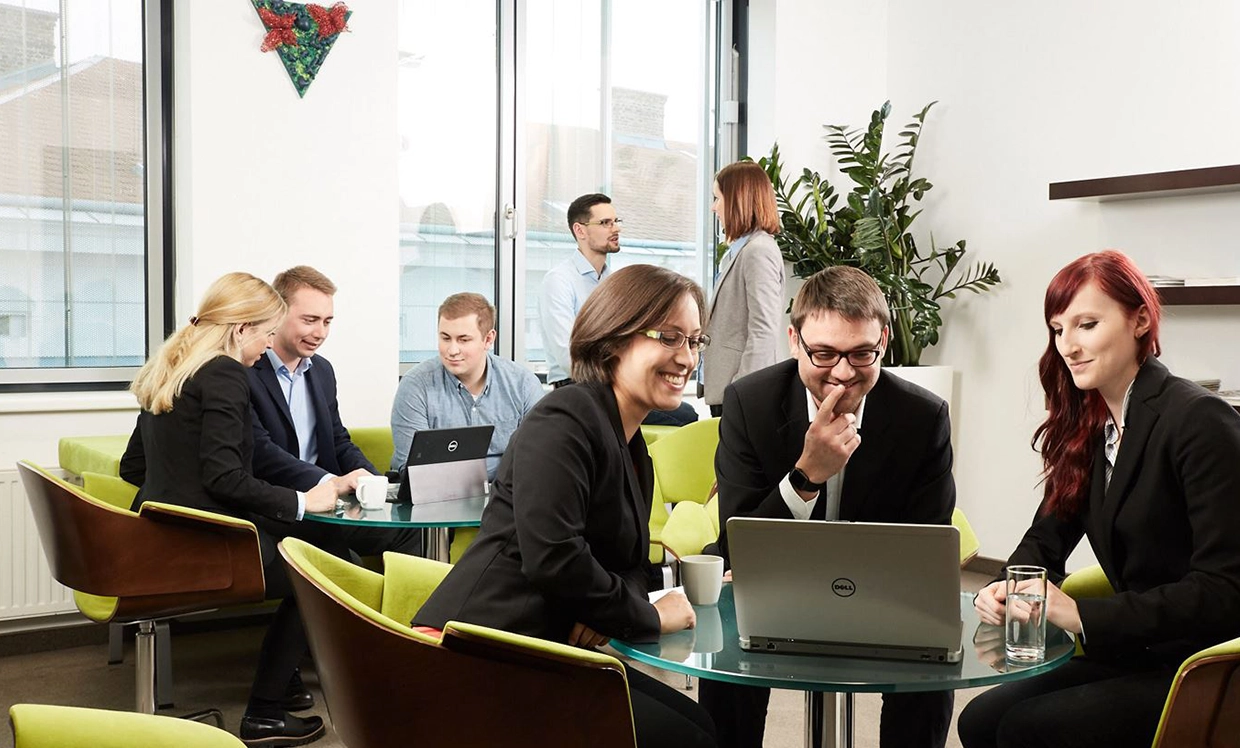 Die VERBUND-Trainees haben sich in Arbeitsgruppen zusammengetan. Wir sehen zwei Dreiergruppen. Sie sind alle im legeren Business-Look gekleidet. Im Hintergrund führen zwei VERBUND-Mitarbeiter ein Gespräch.