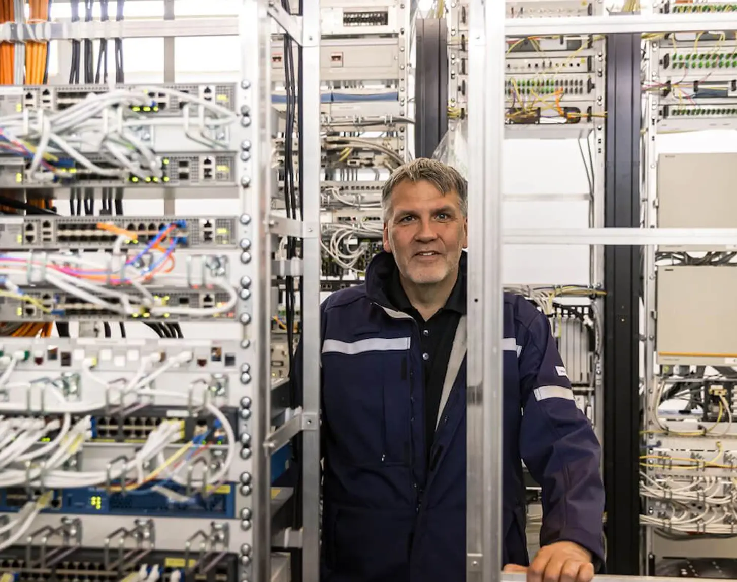 A technical employee with a dark blue security jacket and reflective stripes on his sleeves stands between several steel shelves with servers. He smiles into the camera.