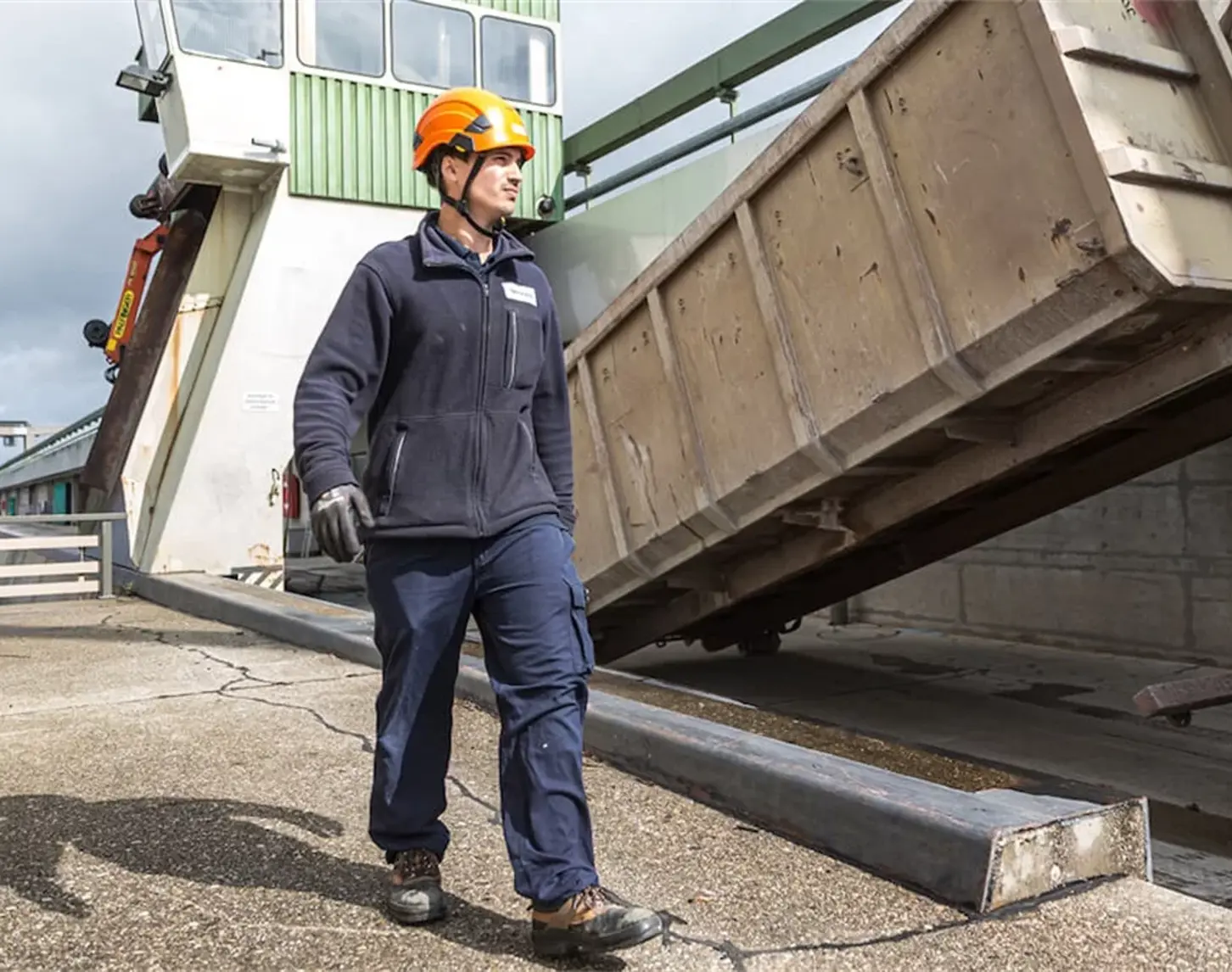 A VERBUND employee in work clothes moves around outdoors at a hydropower plant. The sun is shining. He is wearing an orange-coloured helmet.