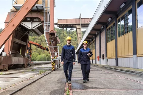 Zwei VERBUND-Mitarbeiter laufen auf die Kamera zu. Beide trägen Arbeitskleidung. Im Hintergrund sehen wir das Wasserkraftwerk Aschach.