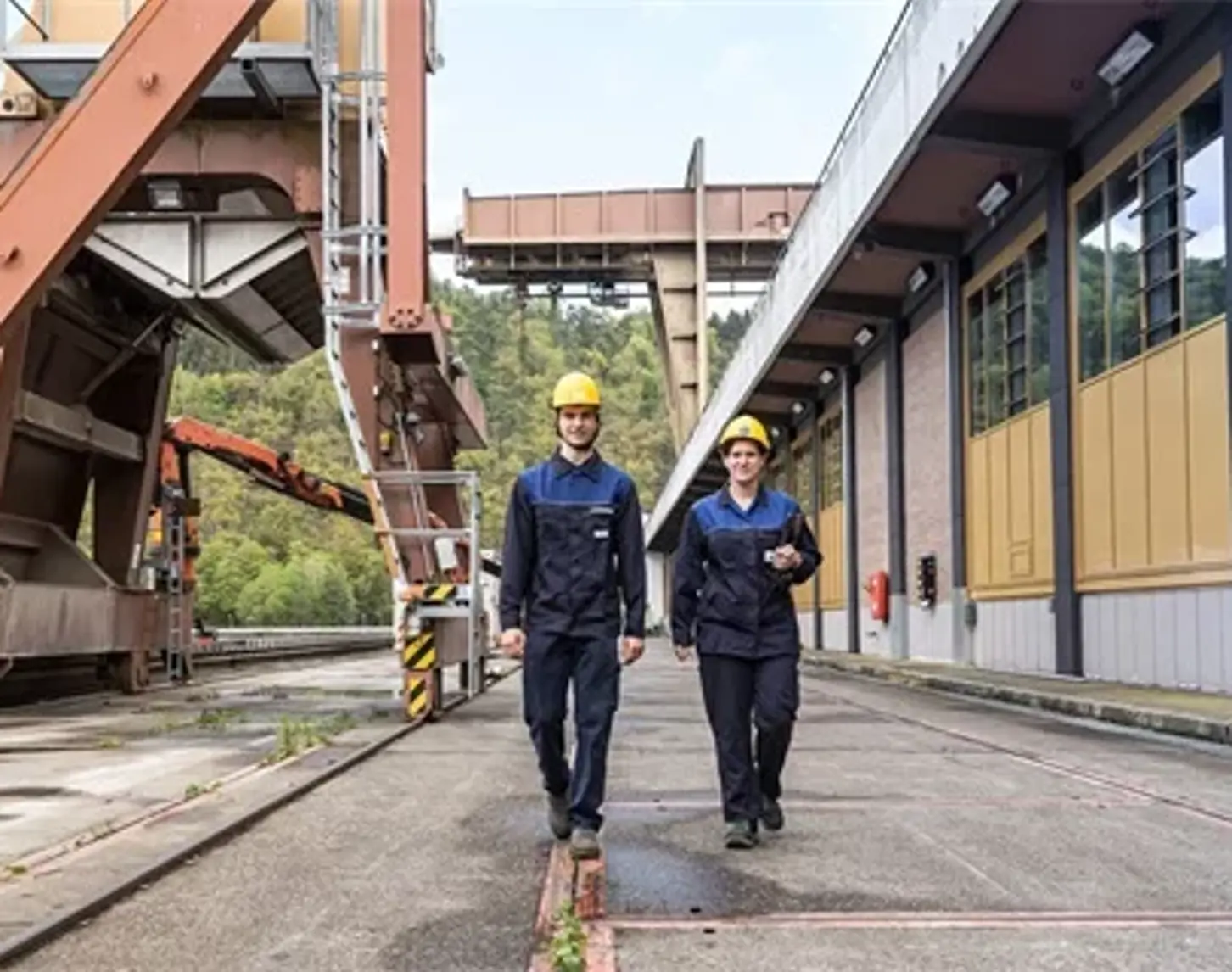 Two VERBUND employees walk towards the camera. Both are wearing work clothes. In the background we see the Aschach hydroelectric power station.