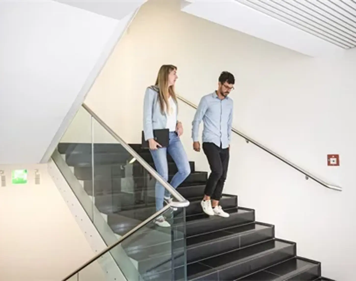 Two people, a man and a woman in business attire, are walking down a staircase, talking animatedly.