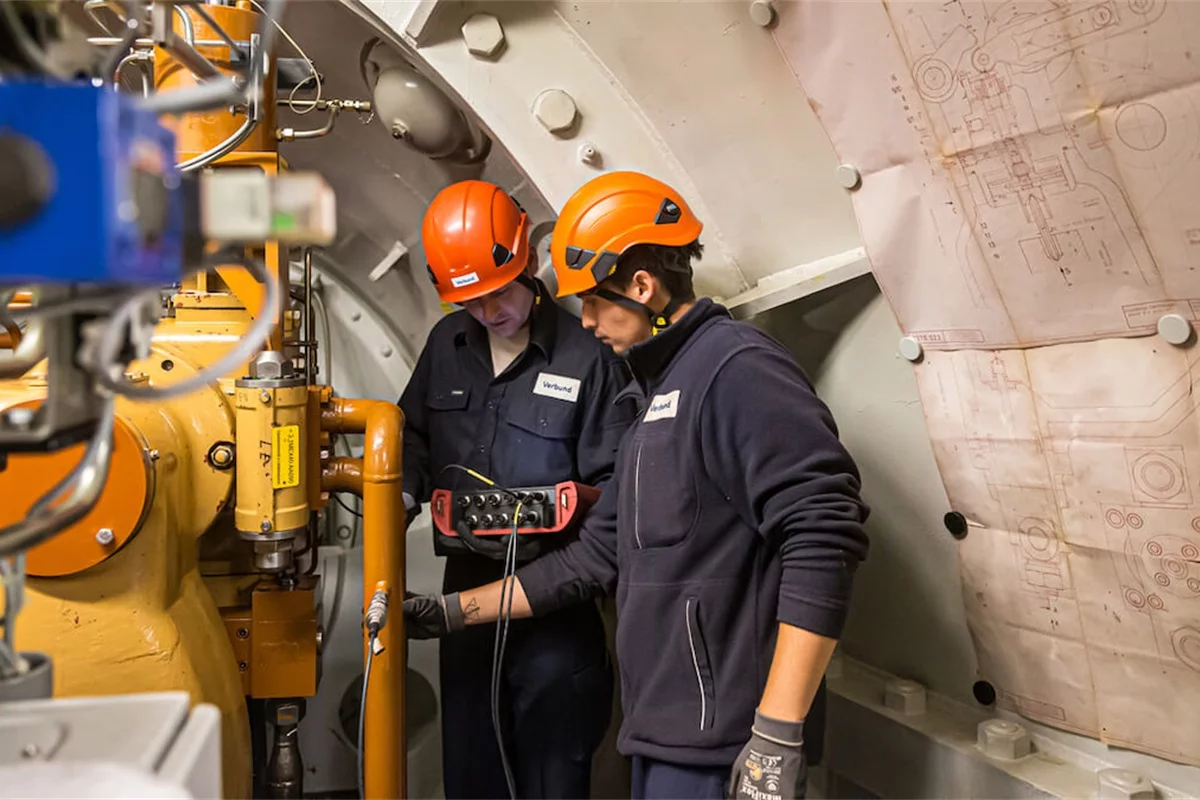 Wir beobachten zwei VERBUND-Mitarbeiter bei ihrer Arbeit im Kraftwerk. Sie tragen dunkelblaue Arbeitskleidung und orangefarbene Helme.