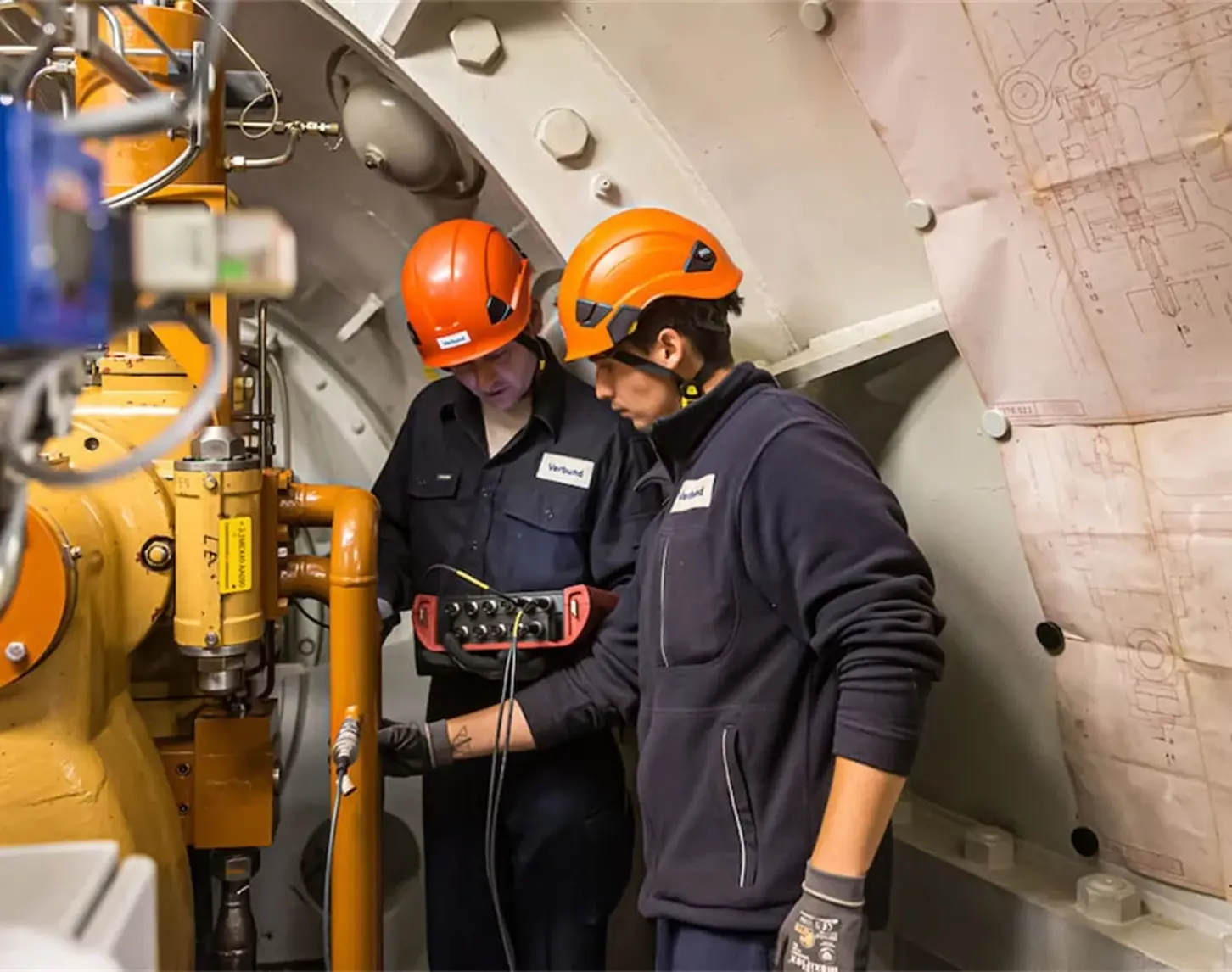 We observe two VERBUND employees at work in the power plant. They are wearing dark blue work clothes and orange helmets.