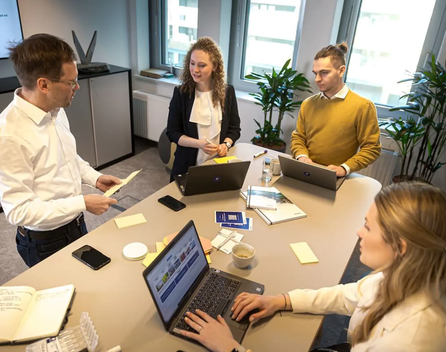 A group of young colleagues are standing around a table. A colleague with a light-coloured blazer and dark blue hair is sitting in front of a laptop, the other colleagues are standing and talking animatedly about a topic.