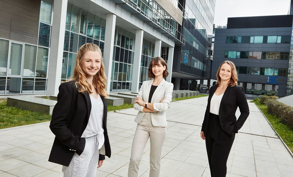 Drei junge Frauen im legeren Business-Look blicken strahlend in die Kamera. Der Hintergrund zeigt größere Gebäude mit vielen Fenstern.