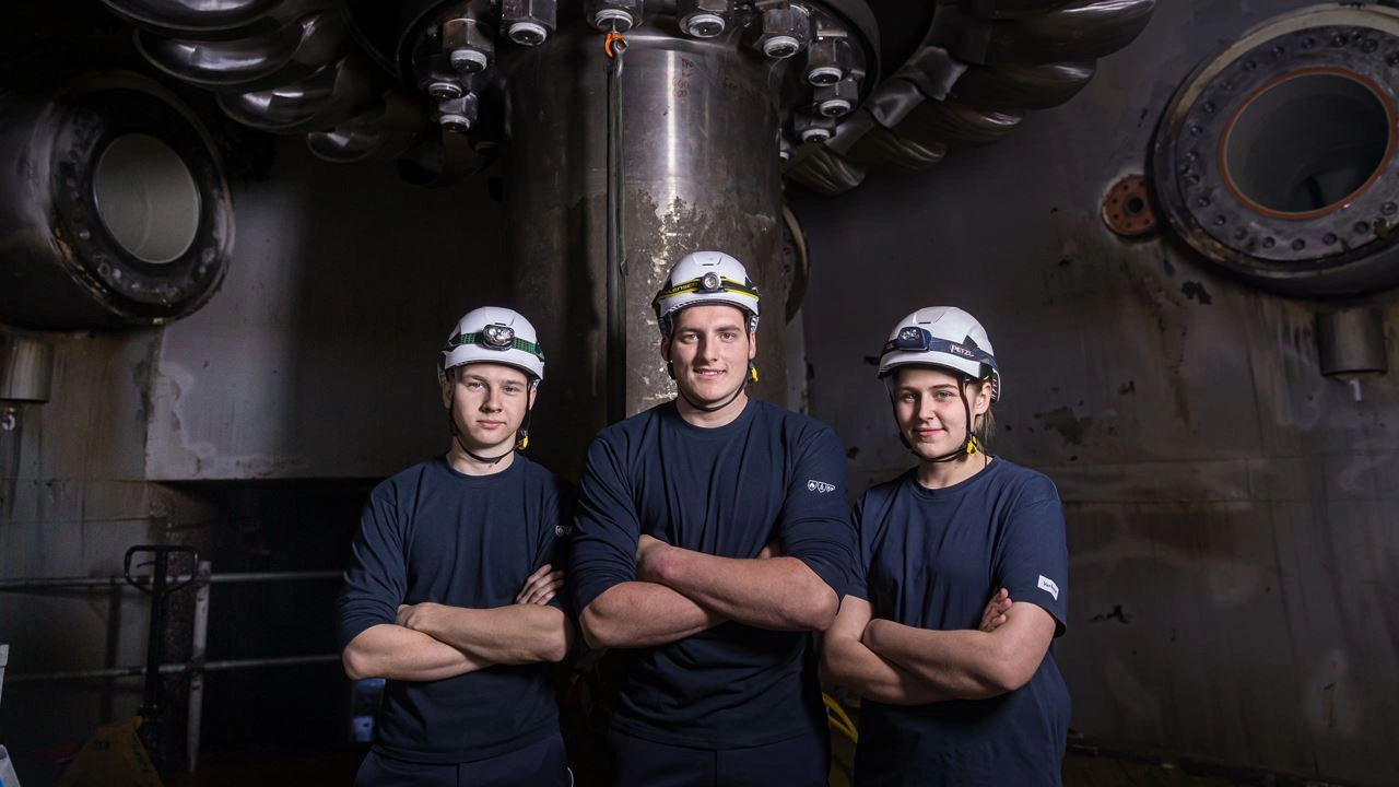 Drei VERBUND-Mitarbeiter:innen posieren im Innen der eines Wasserkrafttwerks. Sie tragen dunkelblaue T-Shirt, weiße Helme. Der Hintergrund ist wenige beleuchtet und daher sehr dunkel.