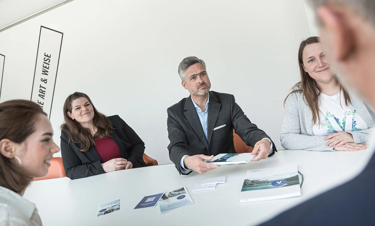 An einem Tisch sitzen 4 Personen von VERBUND und führen ein Bewerbungsgespräch. Drei Damen in Bürokleidung und ein Herr im grauen Anzug sind zu sehen.Die Stimmung ist freundlich und offen. In der Mitte des Tisches liegen Unterlagen zu VERBUND.