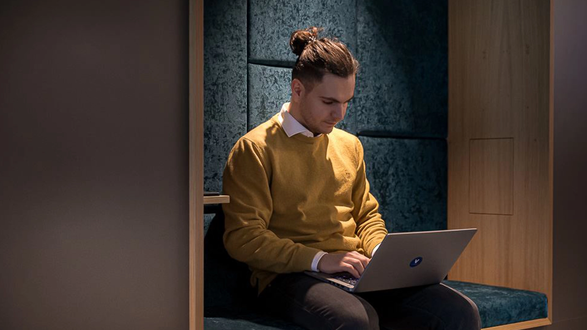 Ein VERBUND-Mitarbeiter sitzt in einer gemütlichen Open-Space-Ecke im Büro. Er trägt sein längeres dunkles Haar in deinen Dutt. Sein Pullover is senffarben und sein Hemd weiß. Sein Laptop hat er auf den Knien. Er tippt.