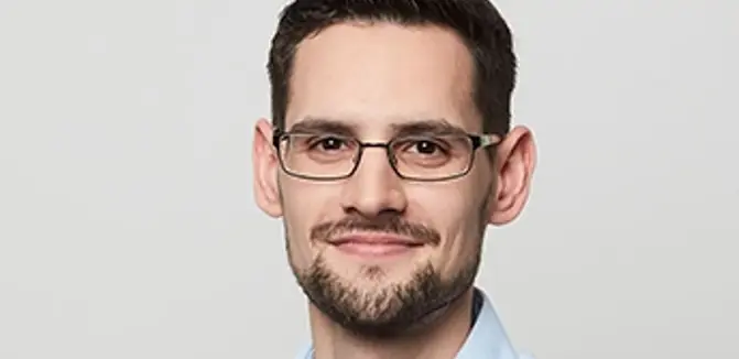 Philip Djakovic hat kurze braune Haare. Er trägt Brille, Bart und hellblaues Hemd.