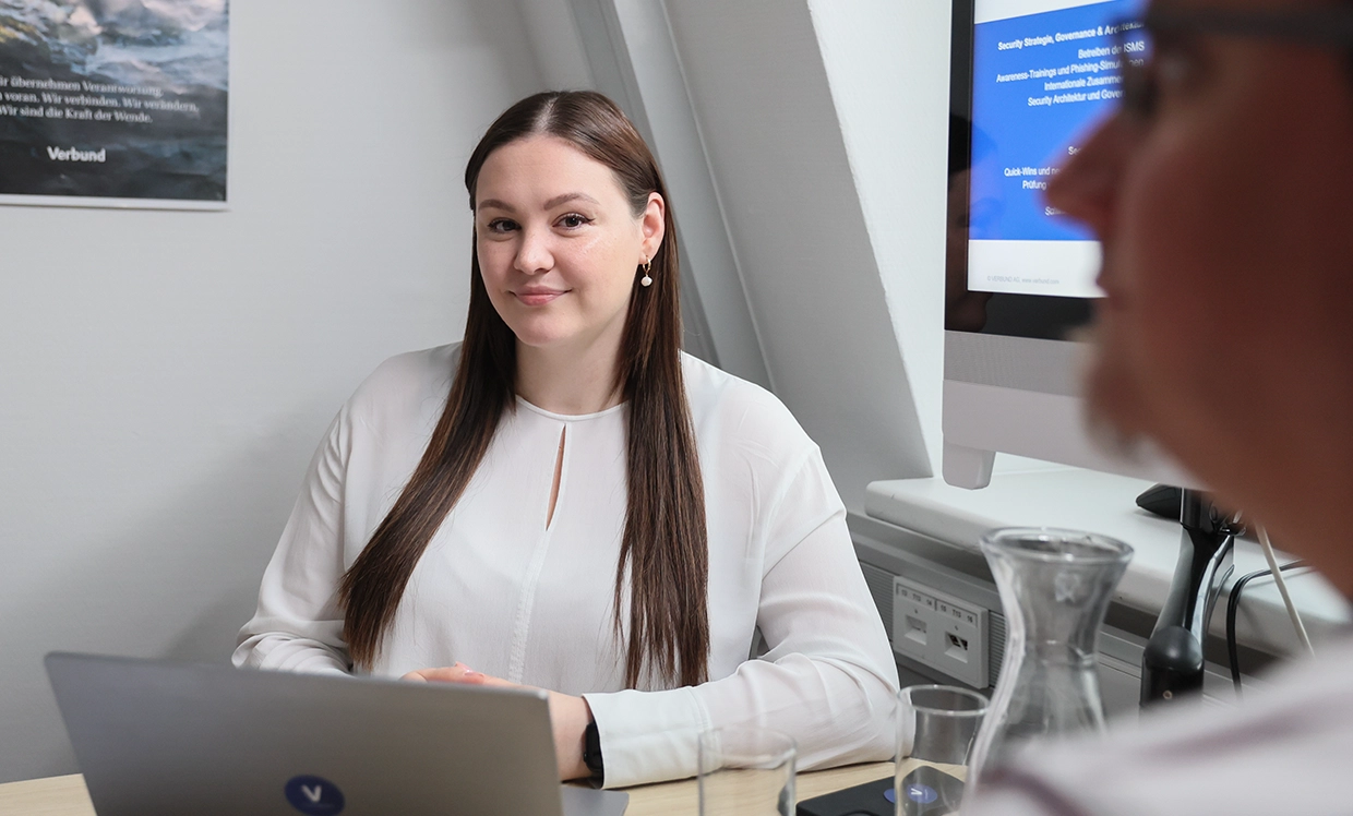 Eine Kollegin mit langen braunen Haaren sitzt vor ihrem Laptop und neben ihr ist eine Präsentation auf einem Bildschirm eingeblendet. Sie lächelt in die Kamera.