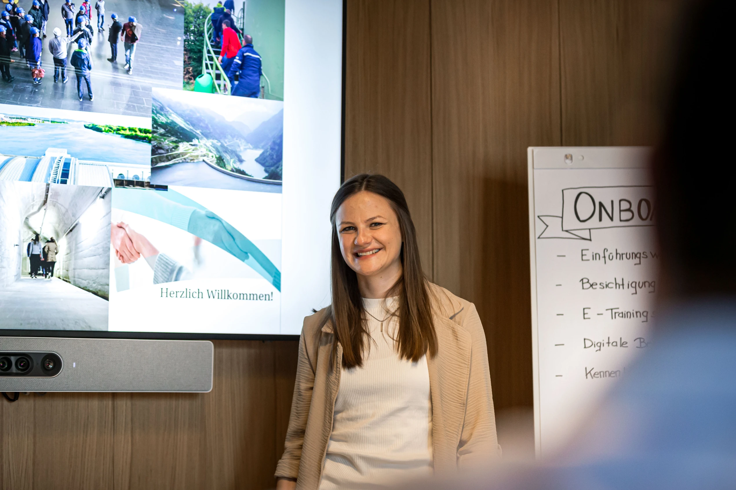 Eine lächelnde Frau steht vor einem Präsentationsbildschirm und einem Flipchart mit der Überschrift „Onboarding“. Der Bildschirm zeigt verschiedene Bilder und den Text „Herzlich Willkommen“. Die Umgebung wirkt professionell und freundlich.