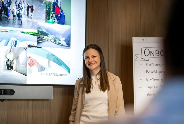 A smiling woman stands in front of a presentation screen and a flip chart with the heading "Onboarding". The screen displays various images and the text "Herzlich Willkommen" (Welcome). The setting appears professional and welcoming.