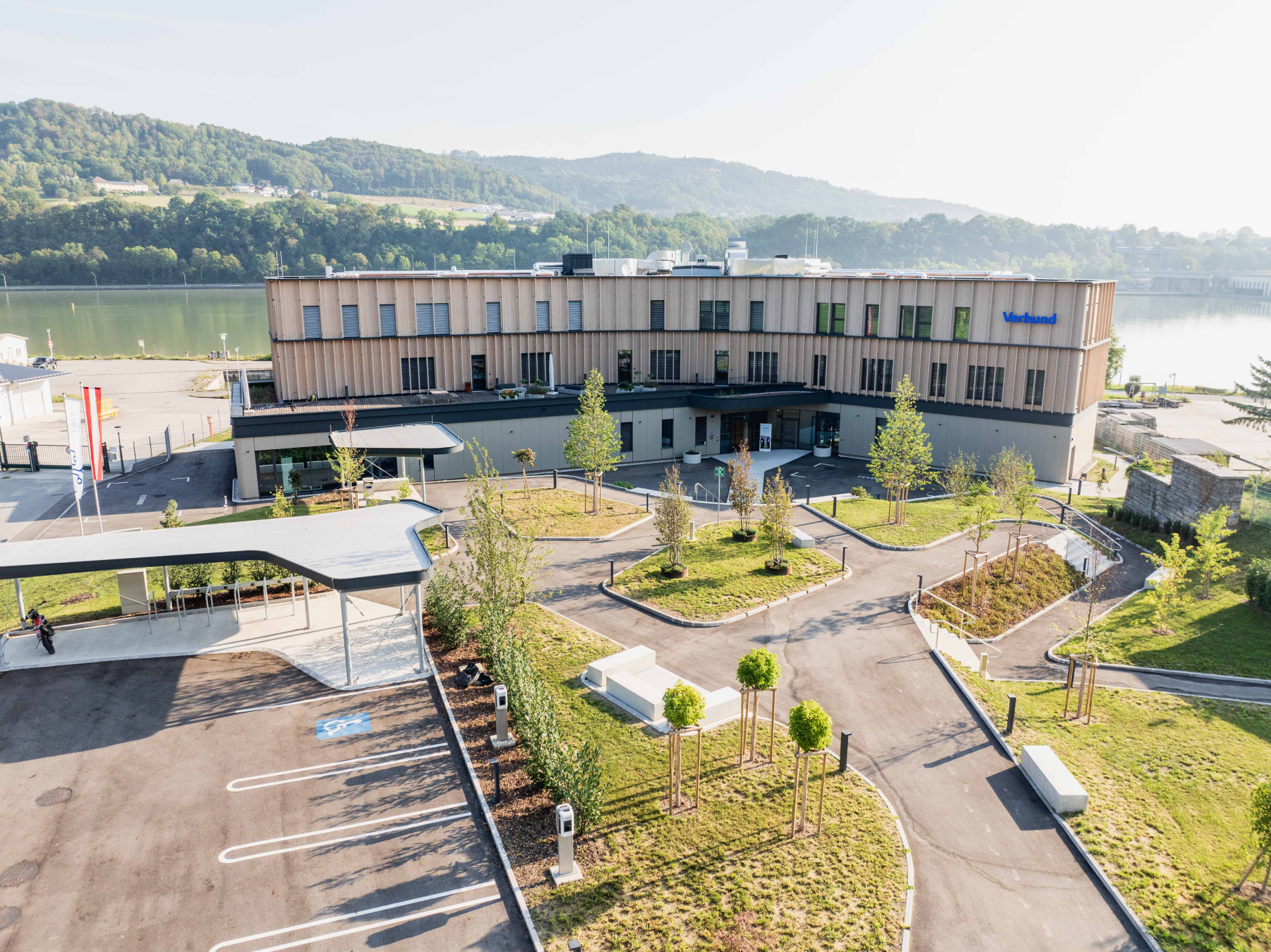 Wir blicken auf das neue Lehrlingsheim im Ybbs. Die Fasade ist beigefarben und blaue VERBUND-Schriftzug ist gut sichtbar. Das Gebäude befindet sich im ländlichen Raum. Im Hintergrund sieht man grüne Hügel.