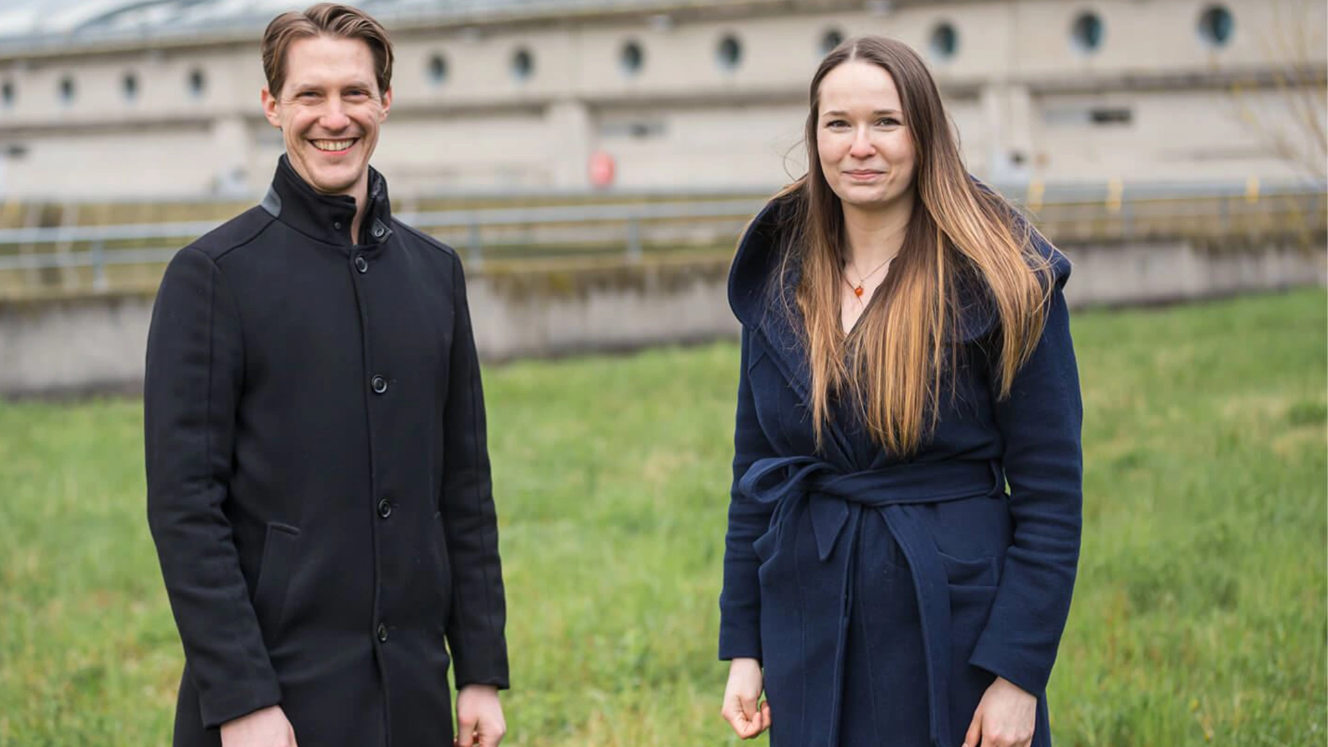 Zwei VERBUND-Kollegen stehen vor einen Waserkraftwerk auf einer grünen Wiese. Die Stimmung ist herbstlich beide tragen leichte Wollmäntel. Er in schwarz und sie in einem schönen dunkelblau.