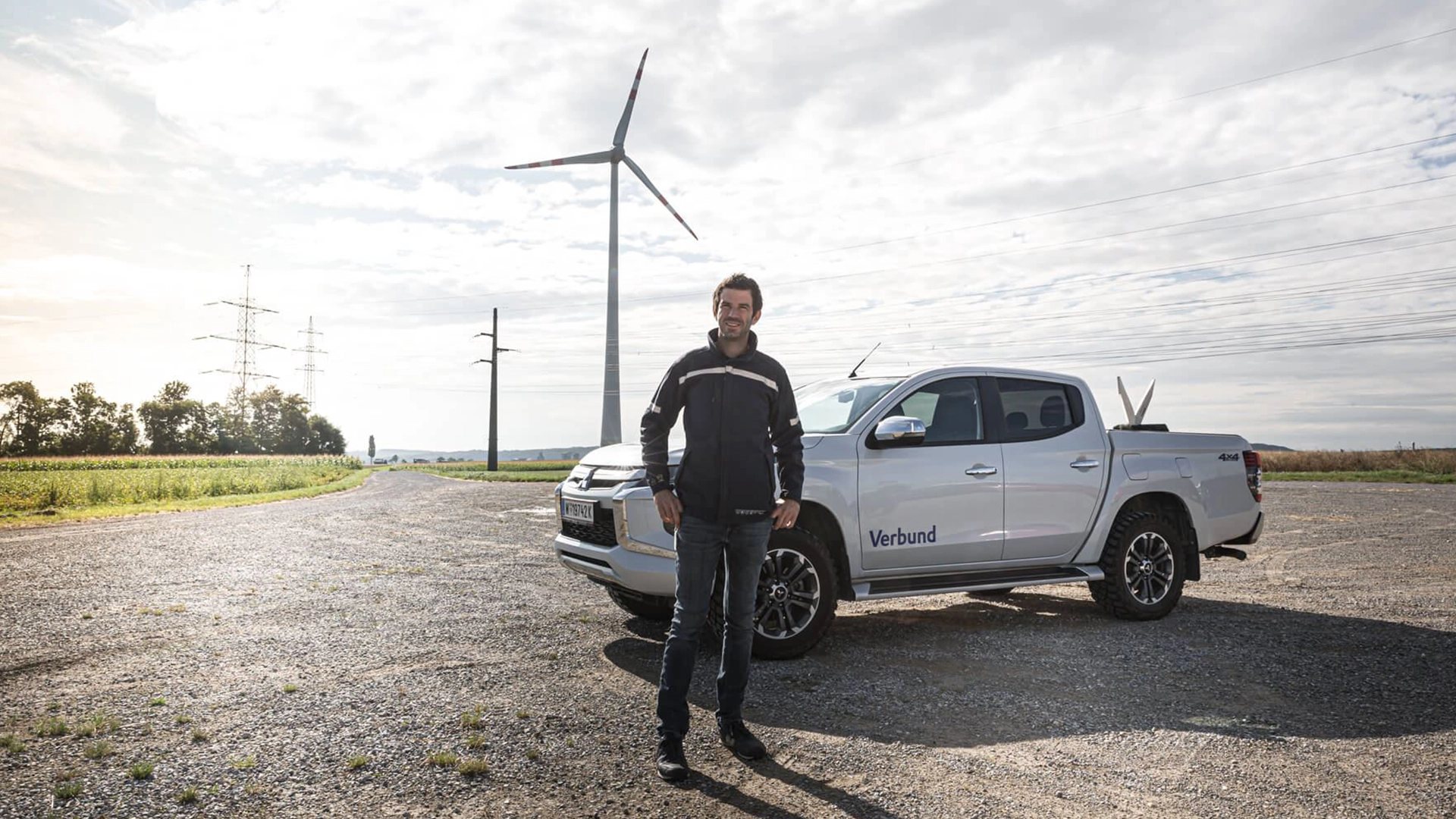 Das Bild zeigt einen VERBUND-Mitarbeiter stehend von einem VERBUND-gebrandeten Auto. Im Hintergrund sehen wir Windränder und Strommasten mit zahlreichen Wolken.