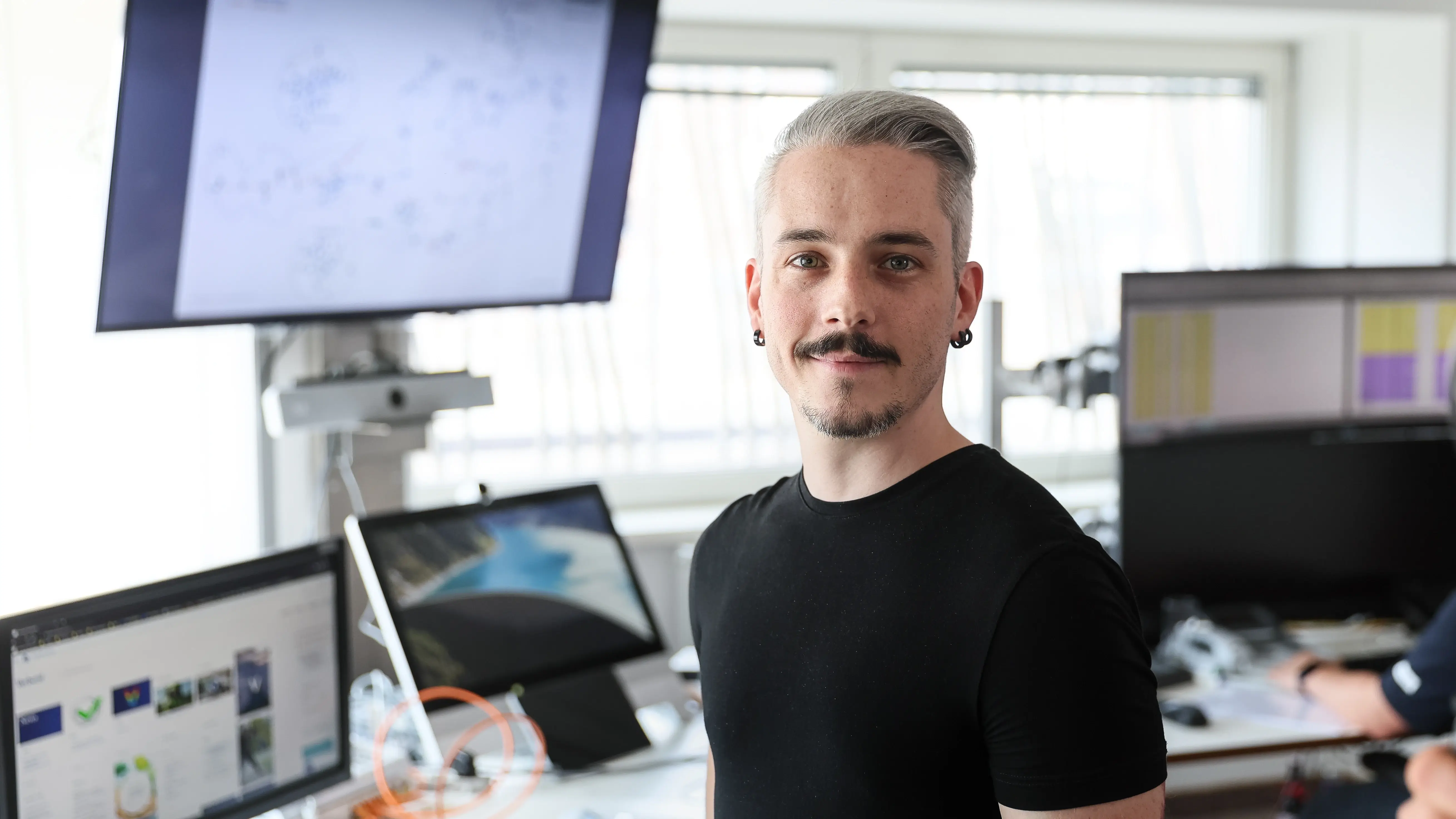 Mann mit dunklem T-Shirt steht in einem hellen Büro vor mehreren Bildschirmen; auf den Monitoren sind Grafiken und Bilder zu sehen.