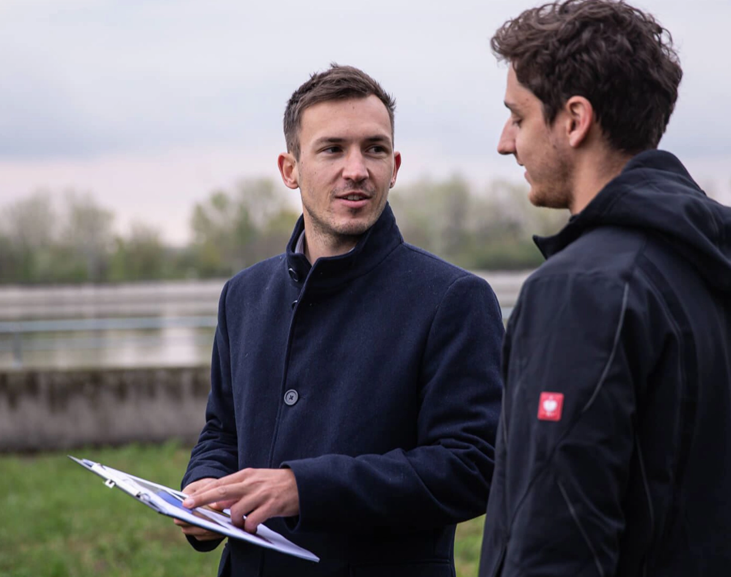 Zwei VERBUND-Mitarbeiter stehen in der Nähe eines Fluss zusammen und unterhalten sich. Einer von beiden hält Dokument in der Hand. Die Stimmung ist herbstlich. Beide tragen Jacken.